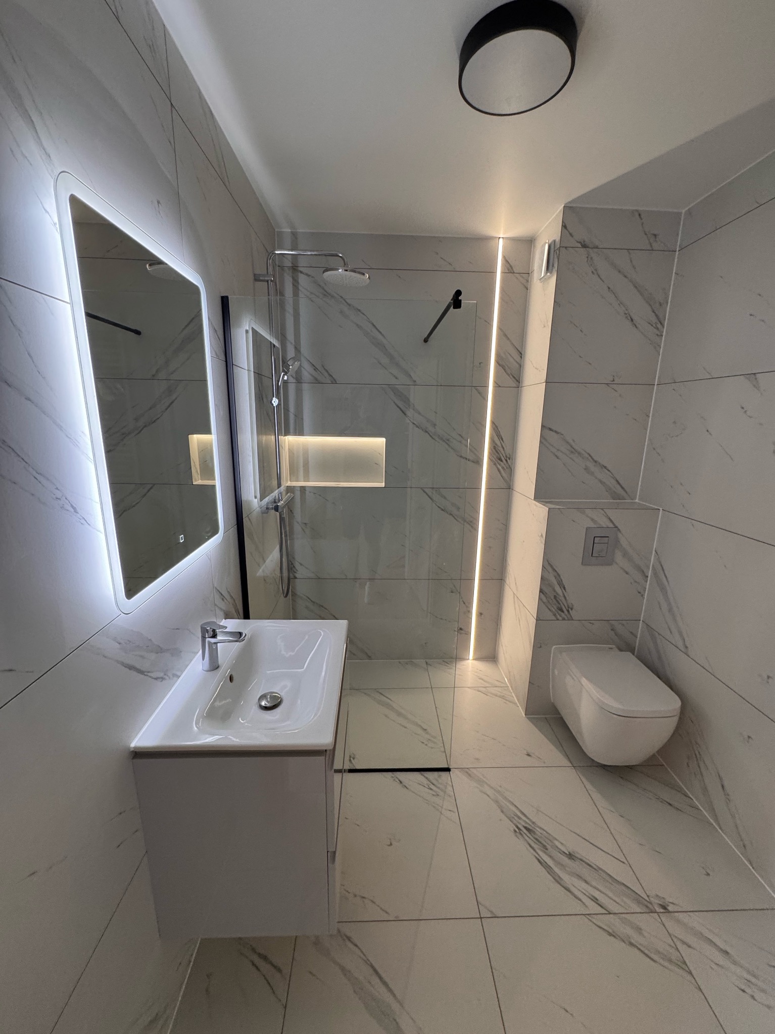Nowoczesna łazienka z marmurowymi płytkami, podświetlanym lustrem, kabiną prysznicową typu walk-in i podtynkową toaletą. Minimalistyczny design, oświetlenie LED.