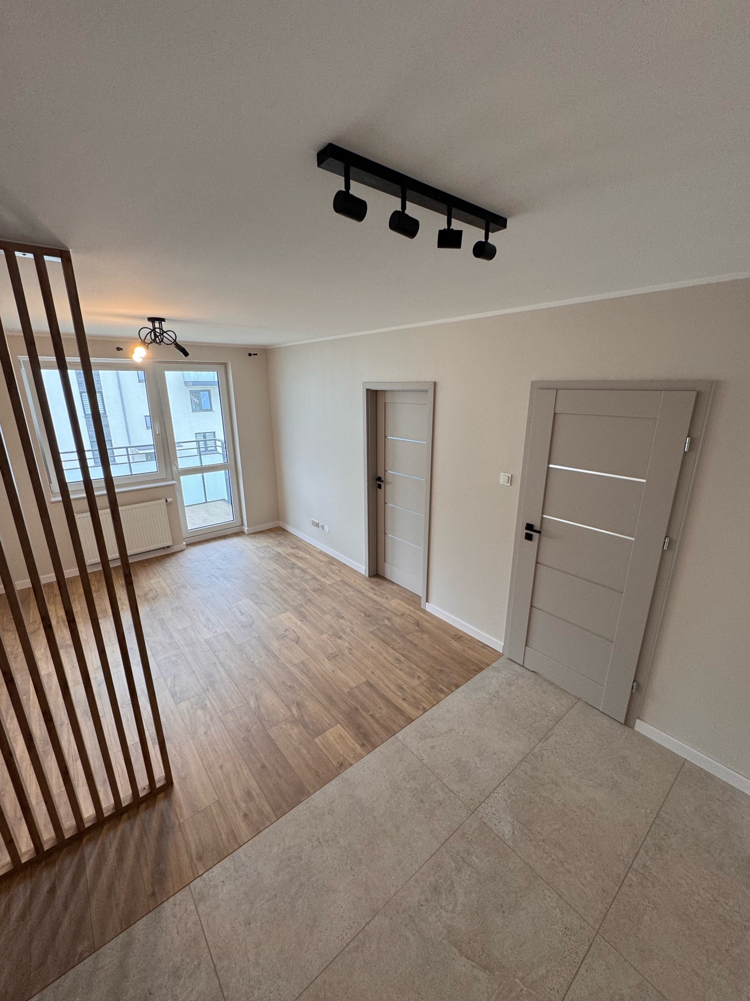 Jasne wnętrze po remoncie: drewnopodobna podłoga, beżowe płytki, dwie szare drzwi z poziomymi wstawkami, drewniana ścianka działowa i oświetlenie sufitowe.