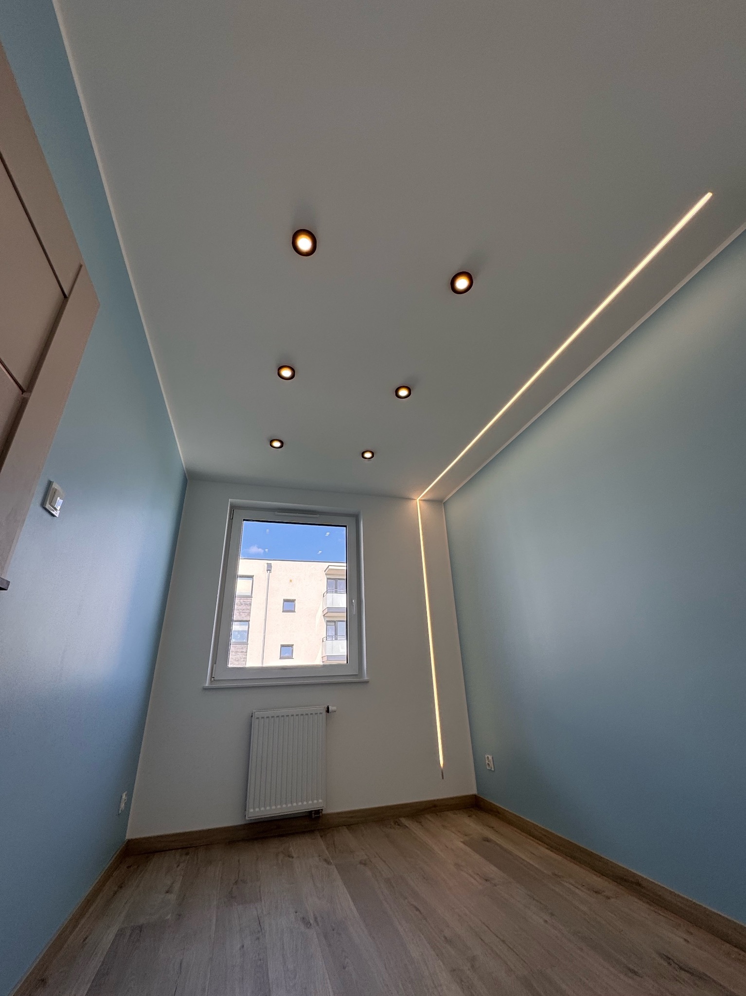 Nowoczesne wnętrze pokoju z błękitnymi ścianami, drewnianą podłogą i oknem. Na suficie zamontowane są punktowe lampy i listwa LED. Widok z dołu, ukazujący detale wykończenia.