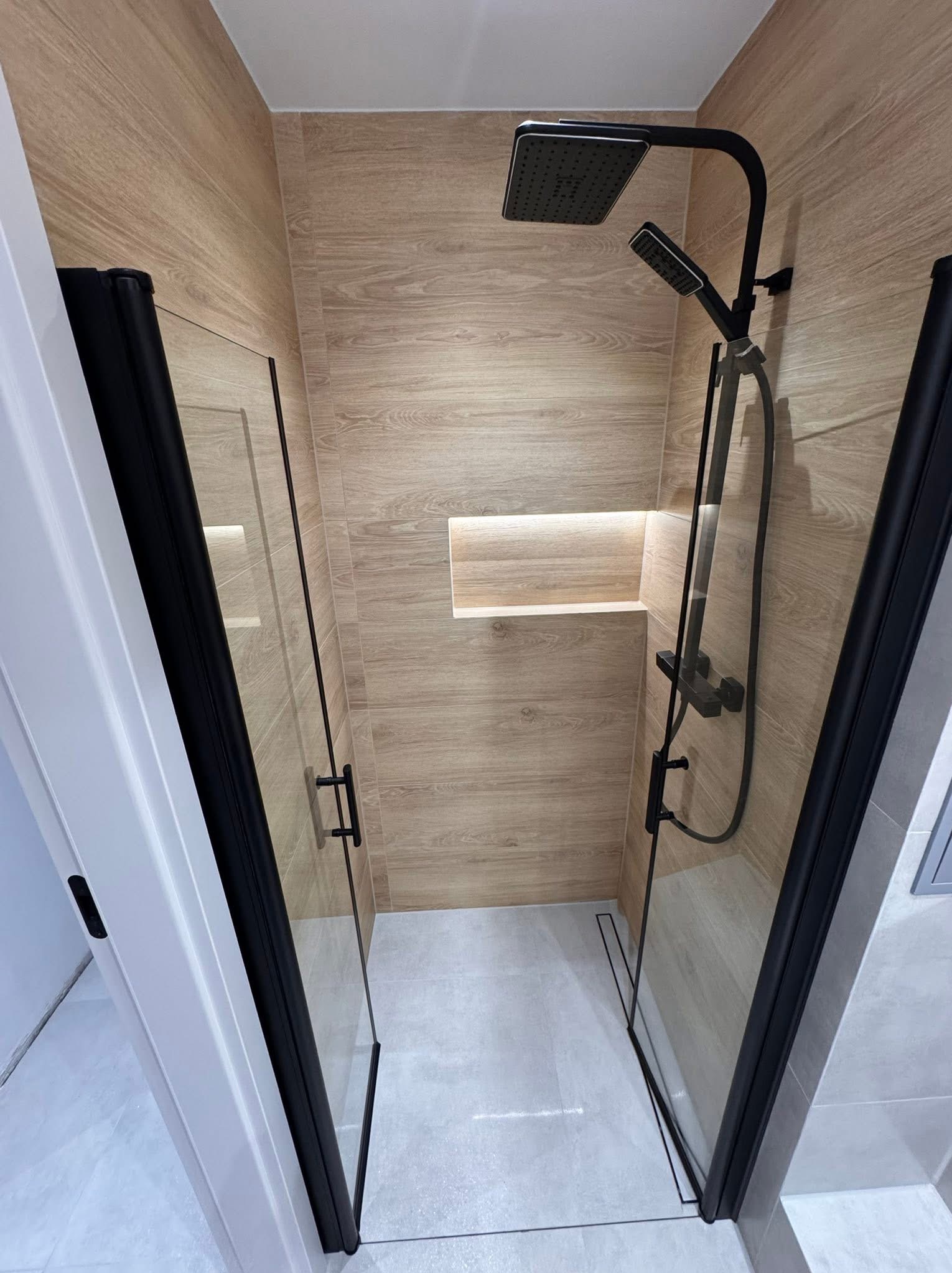 Nowoczesna kabina prysznicowa z czarnymi profilami i deszczownicą, wykończona płytkami imitującymi drewno. W tle wnęka z oświetleniem.