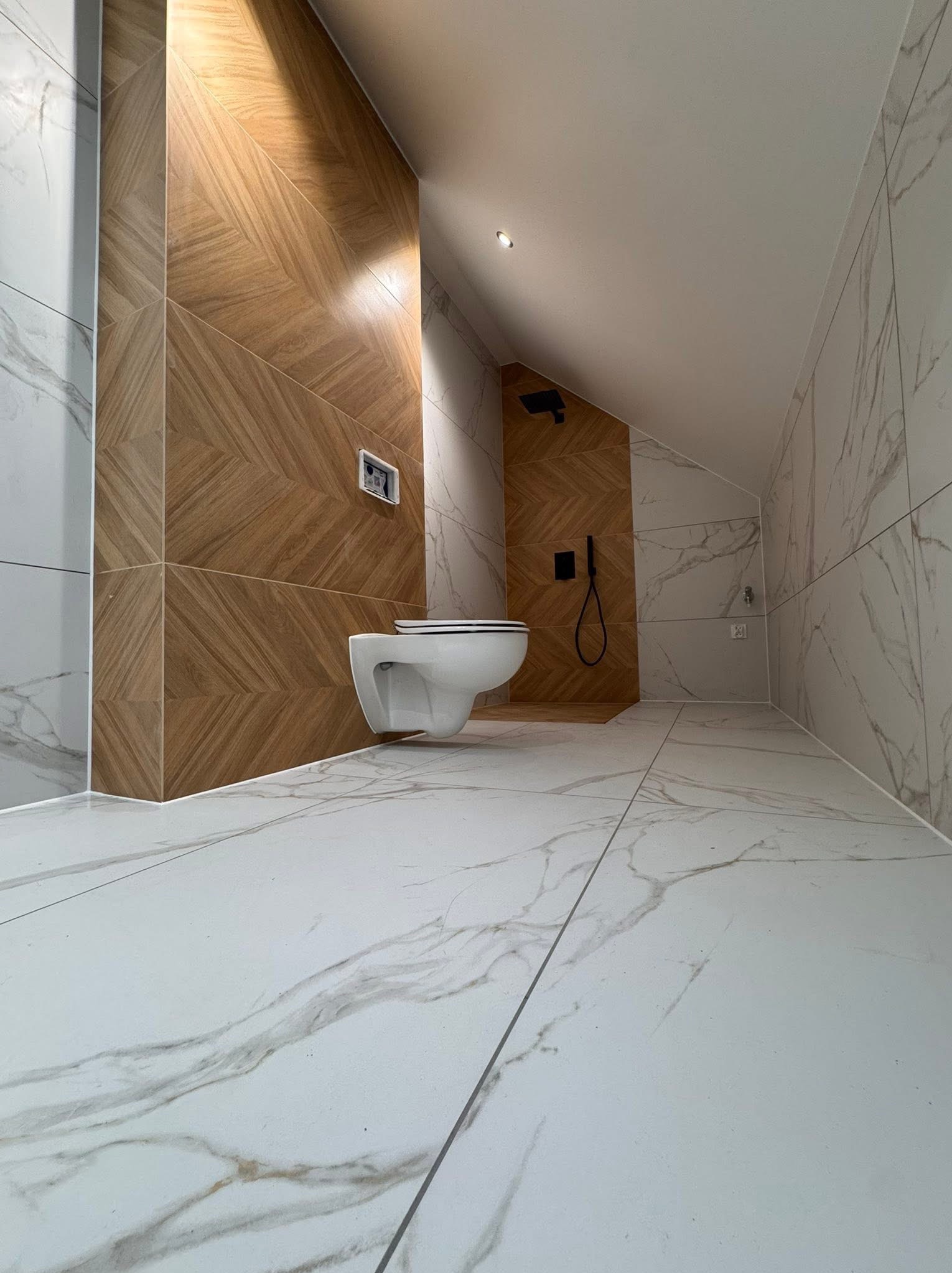 Nowoczesna łazienka na poddaszu z białymi płytkami imitującymi marmur i drewnopodobnymi płytkami na ścianie. Uwagę przykuwa wisząca toaleta i czarna armatura prysznicowa.