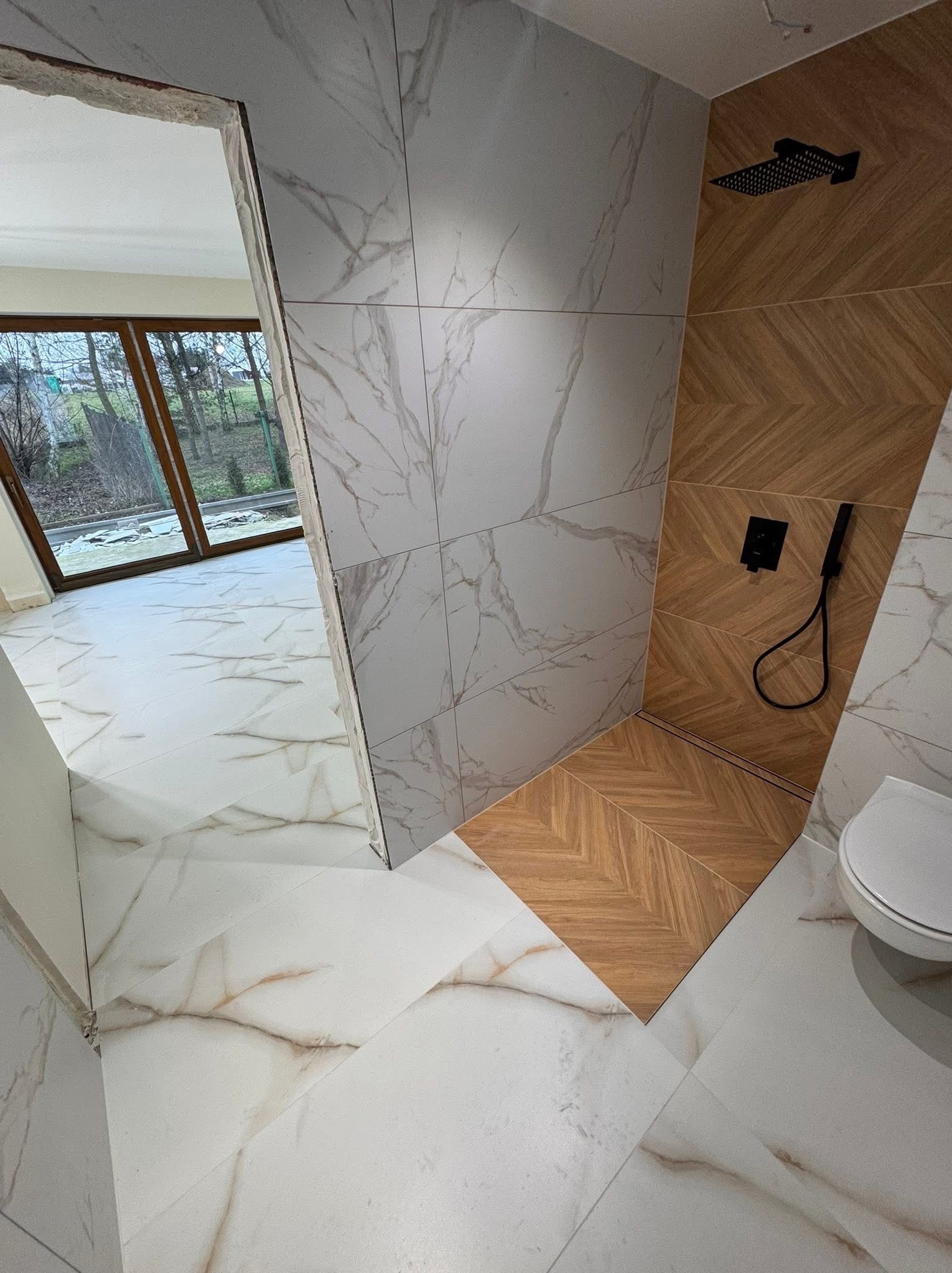 Wykończona łazienka z kabiną typu walk-in. Białe płytki imitujące marmur na ścianach i podłodze, drewnopodobne płytki w strefie prysznica. Czarna armatura.