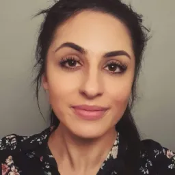 Portret kobiety z ciemnymi włosami upiętymi w kok z ozdobną gumką, ubranej w bluzkę w kwiaty na tle szarej ściany.