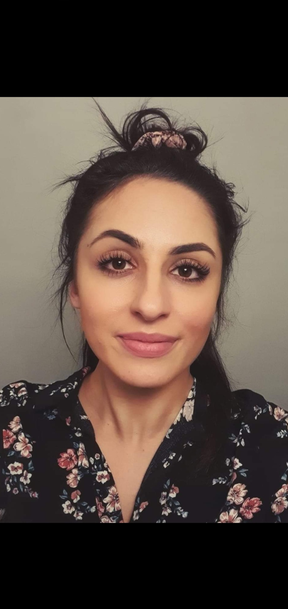 Portret kobiety z ciemnymi włosami upiętymi w kok z ozdobną gumką, ubranej w bluzkę w kwiaty na tle szarej ściany.