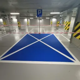Oznakowane niebieskim kolorem miejsce parkingowe dla osoby niepełnosprawnej z białymi liniami na szarej posadzce w garażu podziemnym, zabezpieczone taśmą ostrzegawczą.