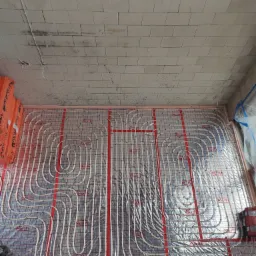 Widok z góry na rozłożoną instalację ogrzewania podłogowego marki Roth na folii aluminiowej z czerwoną siatką, przed wylaniem wylewki, widoczny surowy strop z bloczków betonowych.