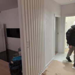 IDAO Ivan Hrynchuk - Nowoczesna ściana z pionowymi panelami w odcieniach bieli, widoczny fragment podłogi zabezpieczonej folią malarską, w tle sprzęt budowlany i fragment telewizora, osoba w kurtce w przejściu.