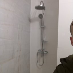 IDAO Ivan Hrynchuk - Prysznic z deszczownicą i panelem natryskowym zamontowany na ścianie wyłożonej marmurkową płytką, widoczne fragment osoby w prawym dolnym rogu.