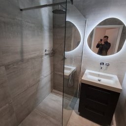 IDAO Ivan Hrynchuk - Nowoczesna łazienka z kabiną typu walk-in, szarą glazurą imitującą kamień, umywalką z szafką i podświetlanym lustrem o nieregularnym kształcie. Widoczny mężczyzna robiący zdjęcie.