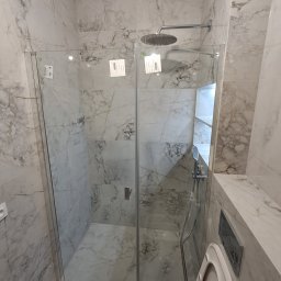 IDAO Ivan Hrynchuk - Szklana kabina prysznicowa walk-in w łazience wykończonej płytami imitującymi marmur w Warszawie. Widoczny panel prysznicowy i toaleta.