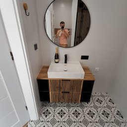 IDAO Ivan Hrynchuk - Nowoczesna łazienka z okrągłym lustrem, białą umywalką na szafce z drewnianymi akcentami i wzorzystą podłogą. Butelka szampana na blacie dodaje elegancji.