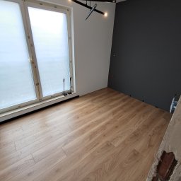 IDAO Ivan Hrynchuk - Jasny pokój po remoncie: drewniana podłoga, biało-grafitowe ściany, nowoczesna lampa sufitowa. Widoczne listwy przypodłogowe przy oknie.