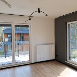 Jasne wnętrze z drewnianą podłogą, białymi ścianami i grzejnikiem. Widoczne duże okna z widokiem na zieleń. Czarna listwa pod sufitem i nowoczesna lampa.