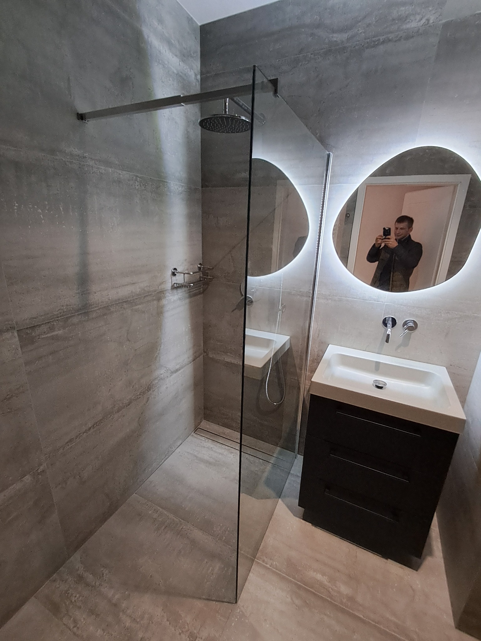 Nowoczesna łazienka z kabiną typu walk-in, szarą glazurą imitującą kamień, umywalką z szafką i podświetlanym lustrem o nieregularnym kształcie. Widoczny mężczyzna robiący zdjęcie.