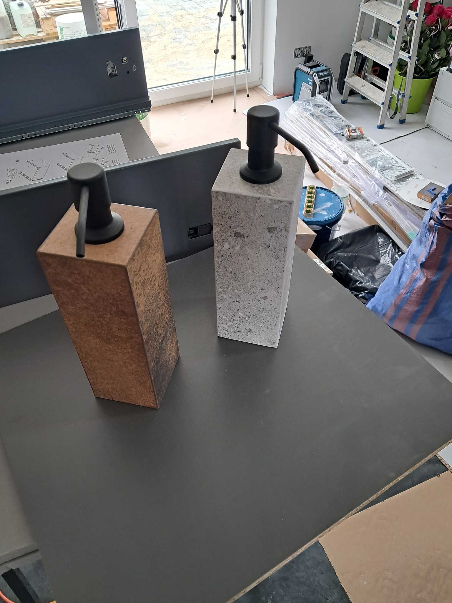 Dwa dozowniki do mydła: betonowy i imitujący drewno, na blacie w trakcie montażu. W tle narzędzia i akcesoria monterskie.