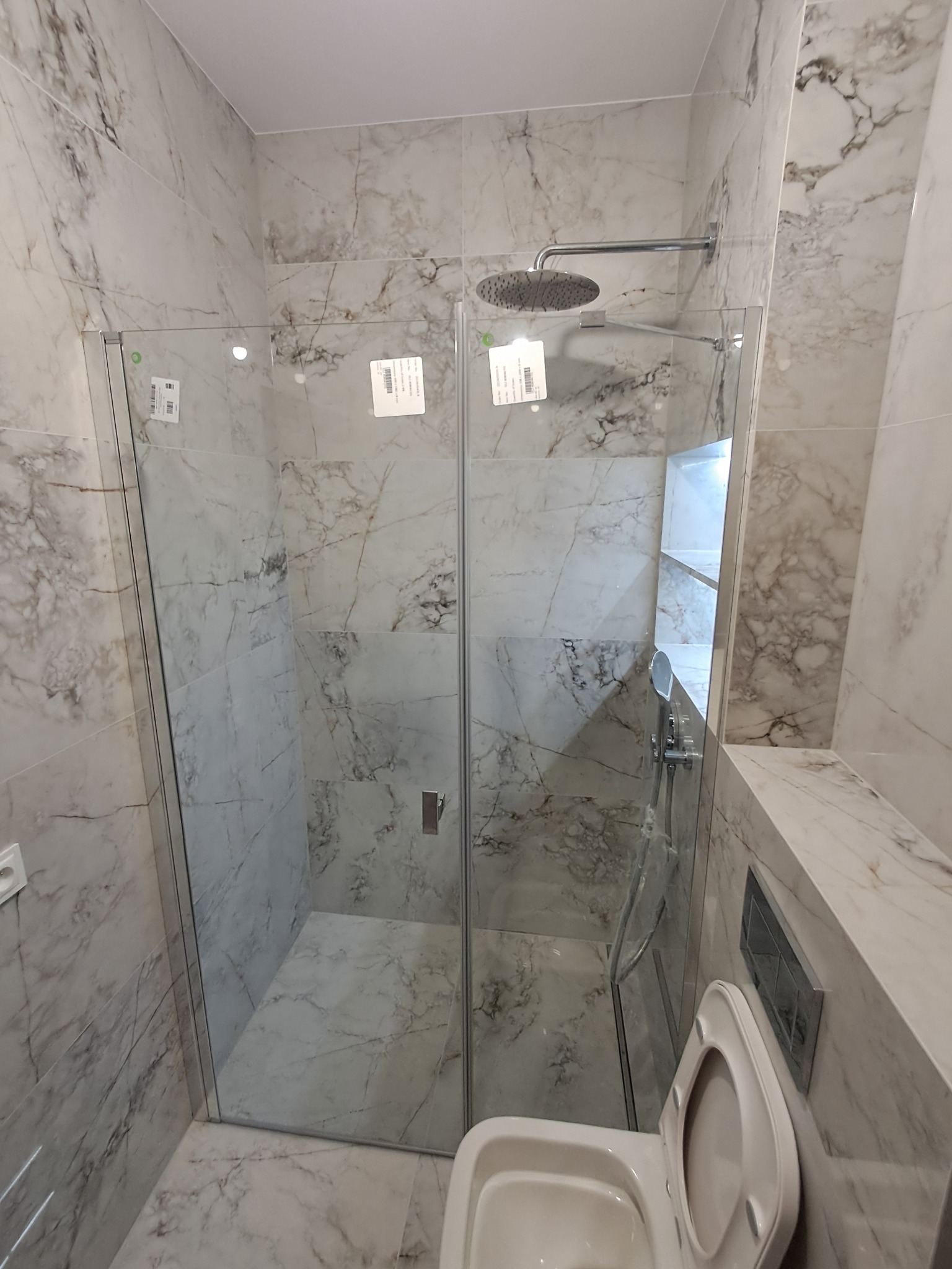 Szklana kabina prysznicowa walk-in w łazience wykończonej płytami imitującymi marmur w Warszawie. Widoczny panel prysznicowy i toaleta.