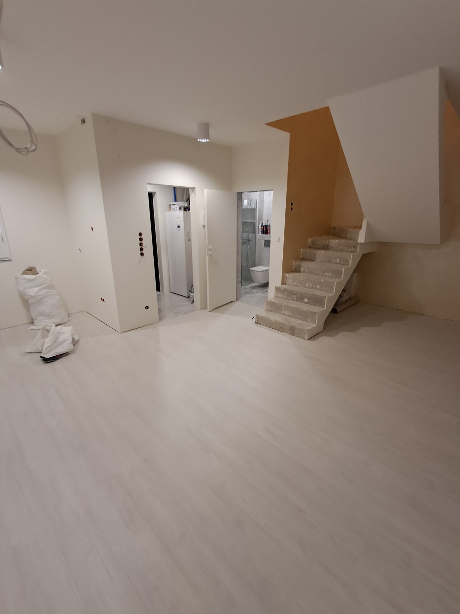 Wykończone, jasne wnętrze z widocznymi schodami w stanie surowym, workami z materiałami budowlanymi i otwartymi drzwiami do łazienki i pomieszczenia gospodarczego.