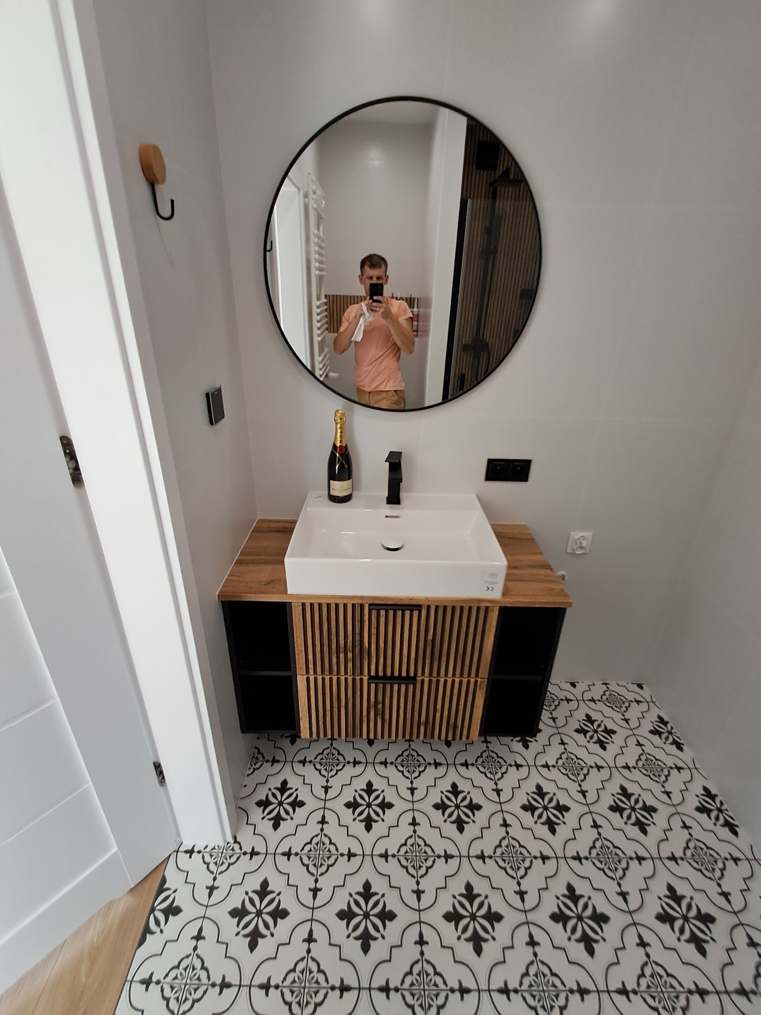 Nowoczesna łazienka z okrągłym lustrem, białą umywalką na szafce z drewnianymi akcentami i wzorzystą podłogą. Butelka szampana na blacie dodaje elegancji.