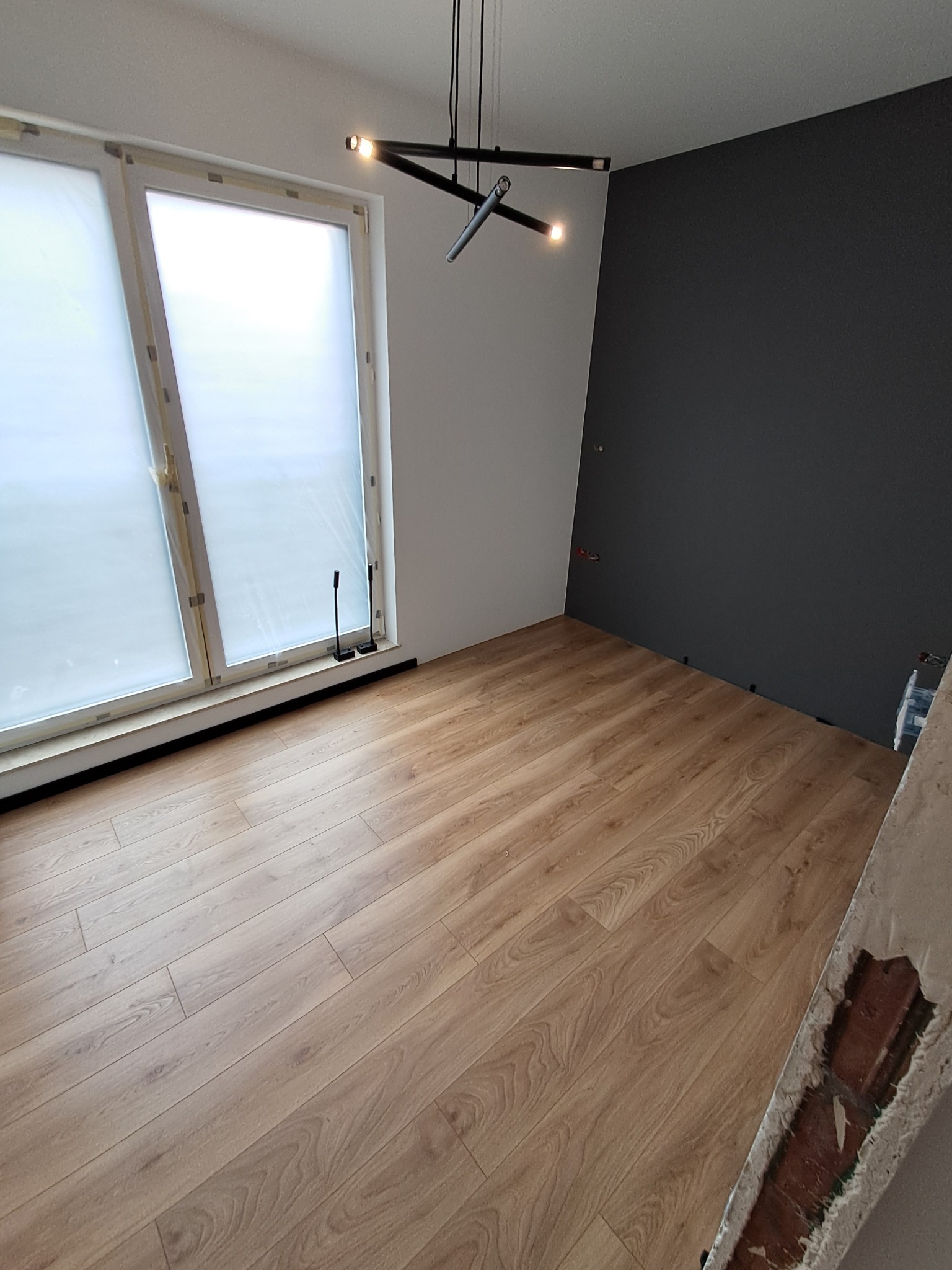 Jasny pokój po remoncie: drewniana podłoga, biało-grafitowe ściany, nowoczesna lampa sufitowa. Widoczne listwy przypodłogowe przy oknie.