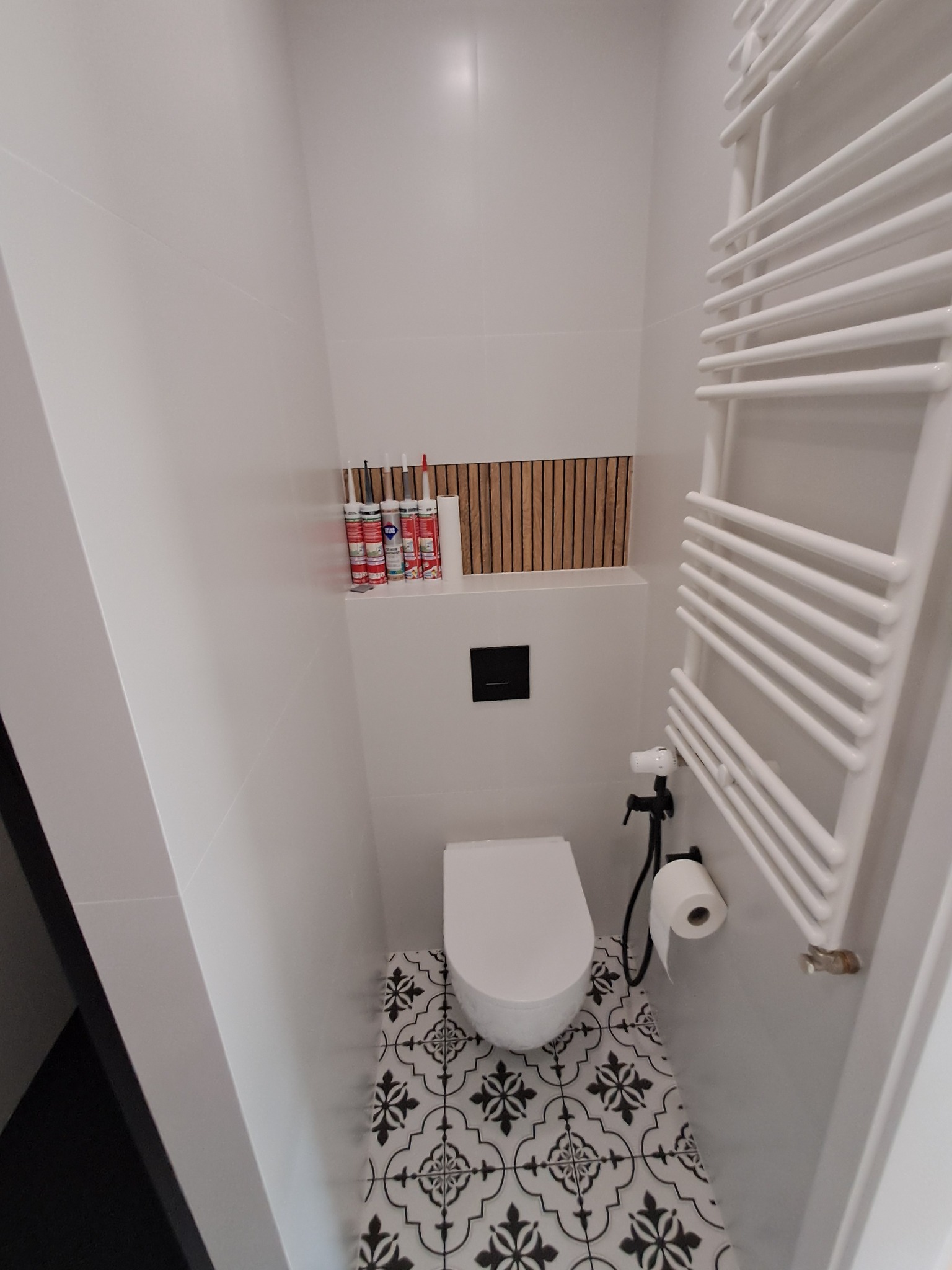Biała toaleta z ozdobną podłogą w wąskiej łazience, grzejnik drabinkowy po prawej, tuby z silikonem na półce. Kontrastowe połączenie wzorów i minimalizmu.