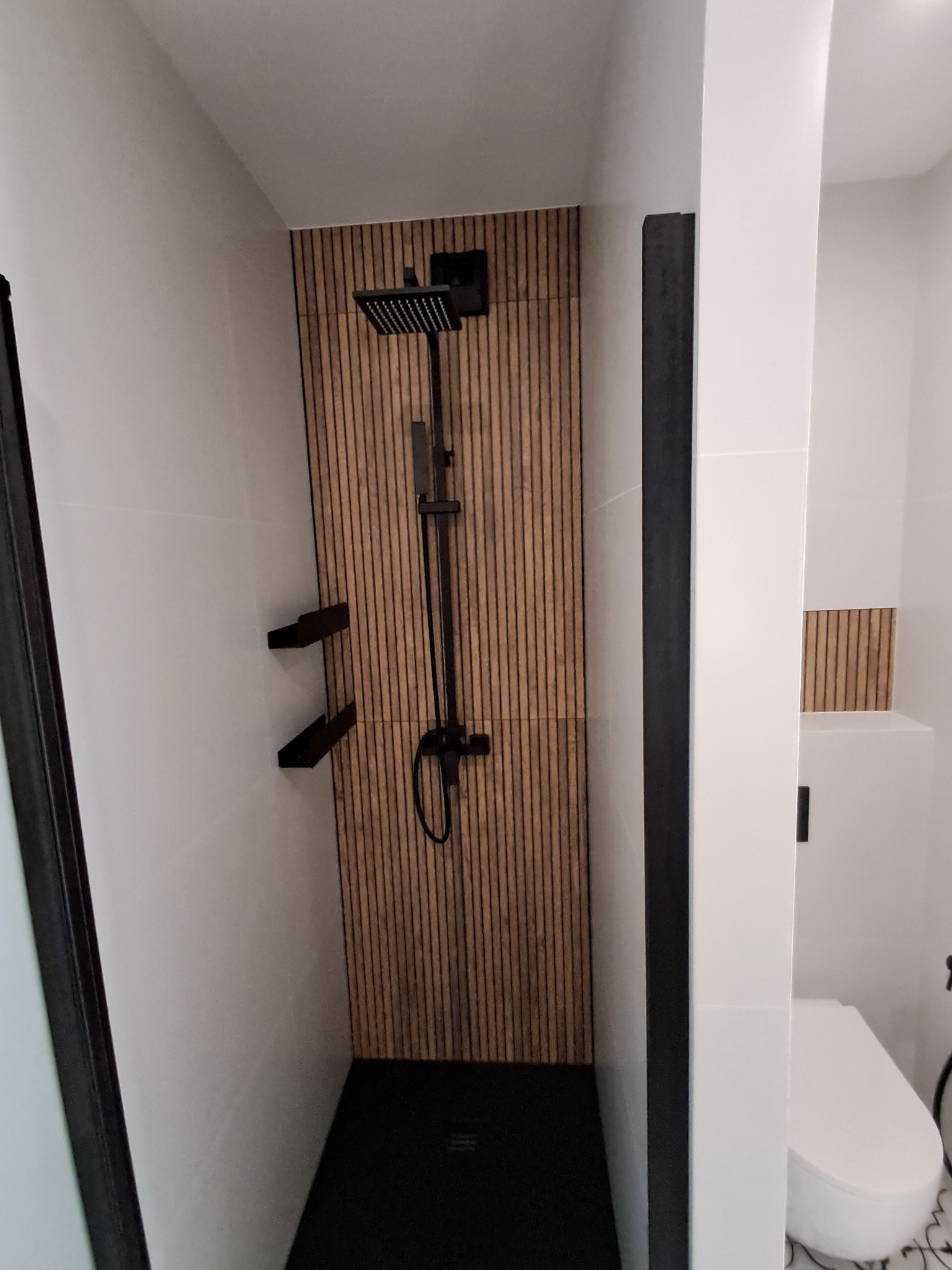 Nowoczesna kabina prysznicowa z czarną armaturą i panelami ściennymi imitującymi drewno, w minimalistycznej łazience z białymi płytkami.