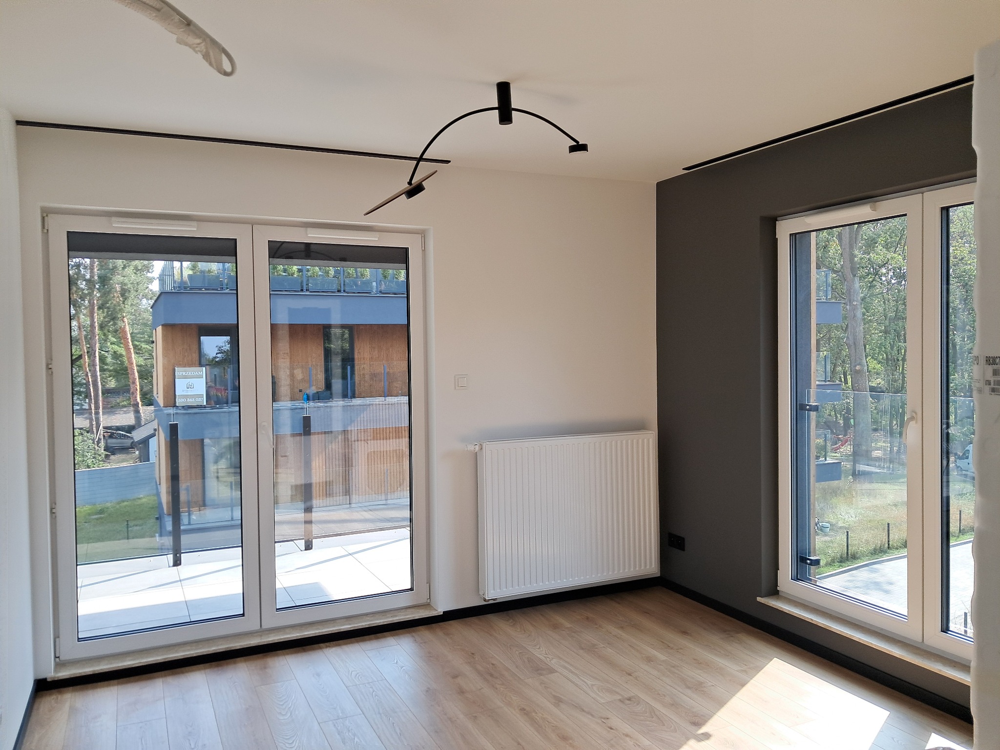 Jasne wnętrze z drewnianą podłogą, białymi ścianami i grzejnikiem. Widoczne duże okna z widokiem na zieleń. Czarna listwa pod sufitem i nowoczesna lampa.