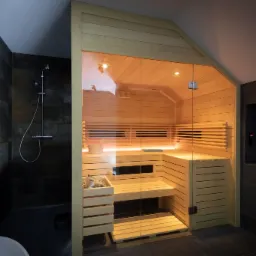 Nowoczesna drewniana kabina z przeszklonymi drzwiami, zlokalizowana w łazience z ciemnymi płytkami i prysznicem, wnętrze oświetlone ciepłym światłem.