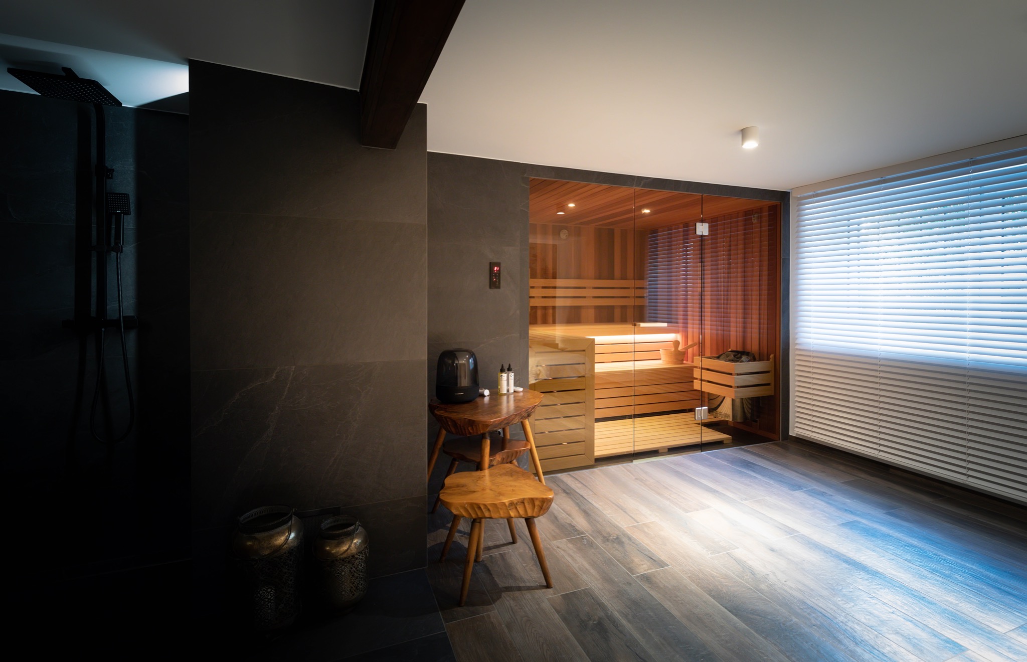 Nowoczesne wnętrze z przeszkloną kabiną, drewnianą ławą w środku i akcesoriami, widoczne designerskie krzesła i stolik z akcesoriami do aromaterapii, całość w stonowanej kolorystyce z elementami...