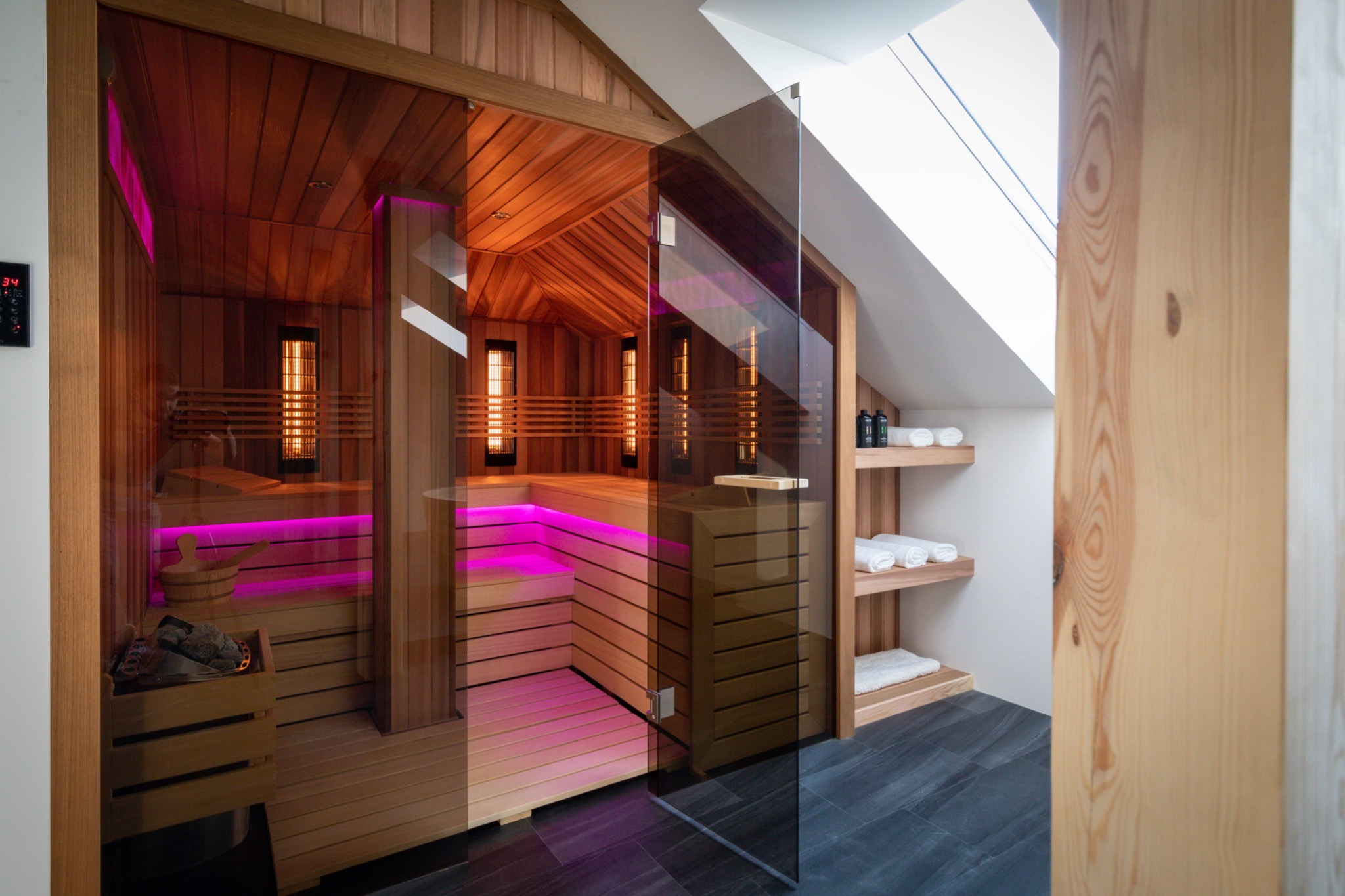Nowoczesna kabina z drewna, ze szklanymi drzwiami i oświetleniem LED w kolorze różowym, widok na wnętrze z grzałkami i akcesoriami do relaksu, drewniane półki z ręcznikami w tle.