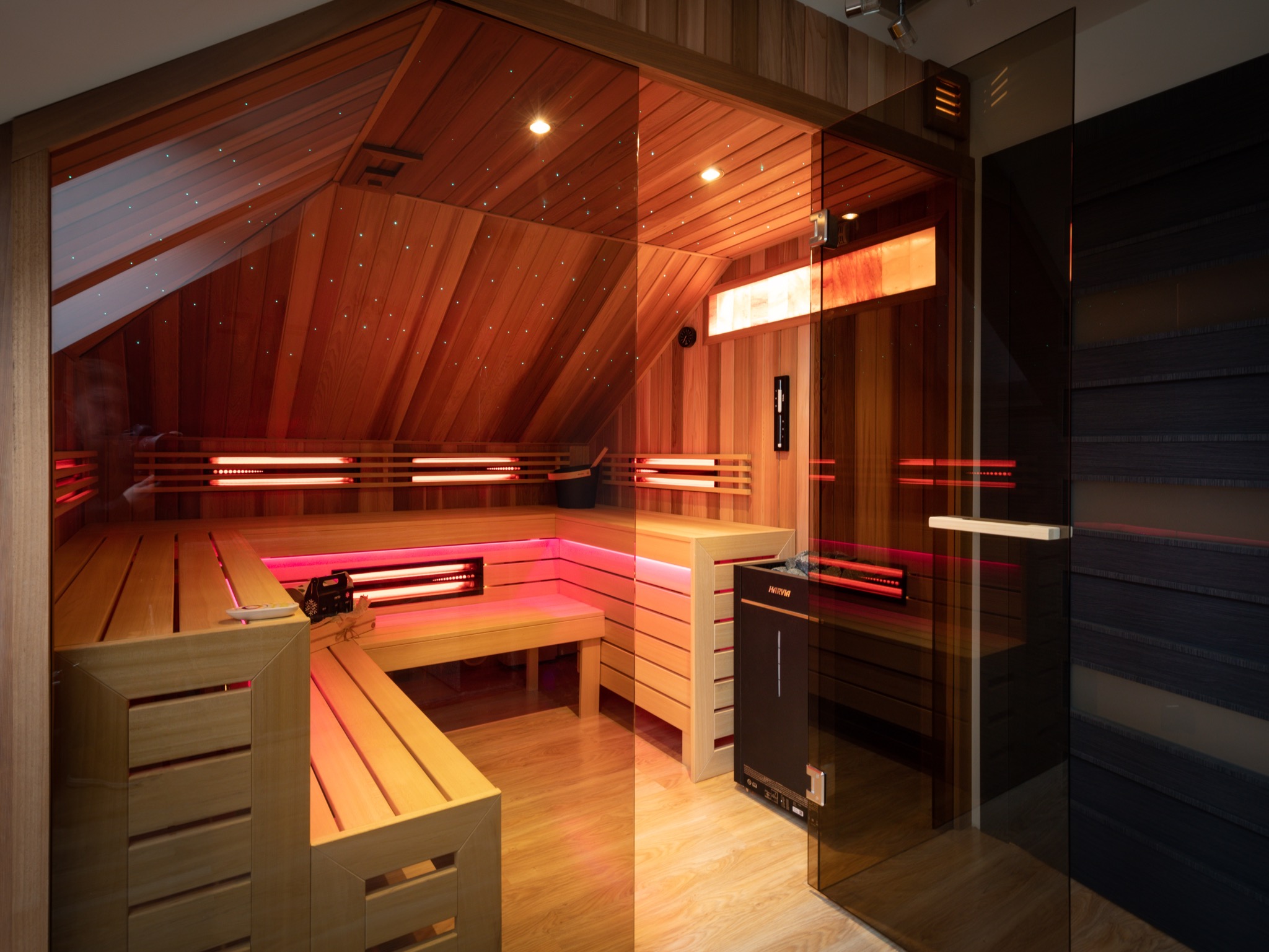Nowoczesna sauna z drewnianym wykończeniem, podświetleniem LED w kolorze czerwonym, imitacją gwiazd na suficie i szklanymi drzwiami, widoczny piec Harvia.