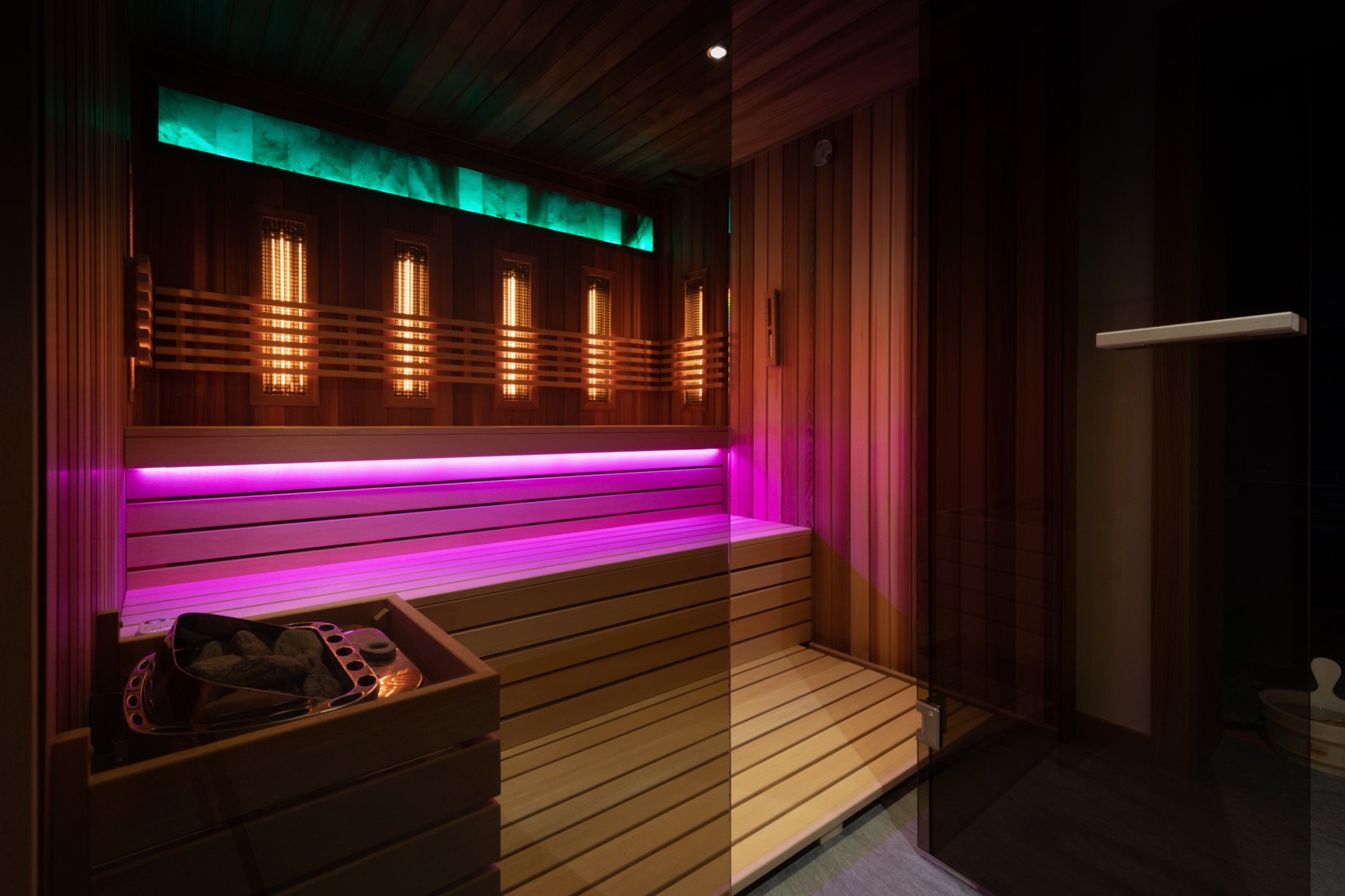 Nowoczesna kabina z drewna z podświetleniem LED w kolorach zielonym i fioletowym, z kamieniami do podgrzewania w drewnianym pojemniku i przeszklonymi drzwiami.