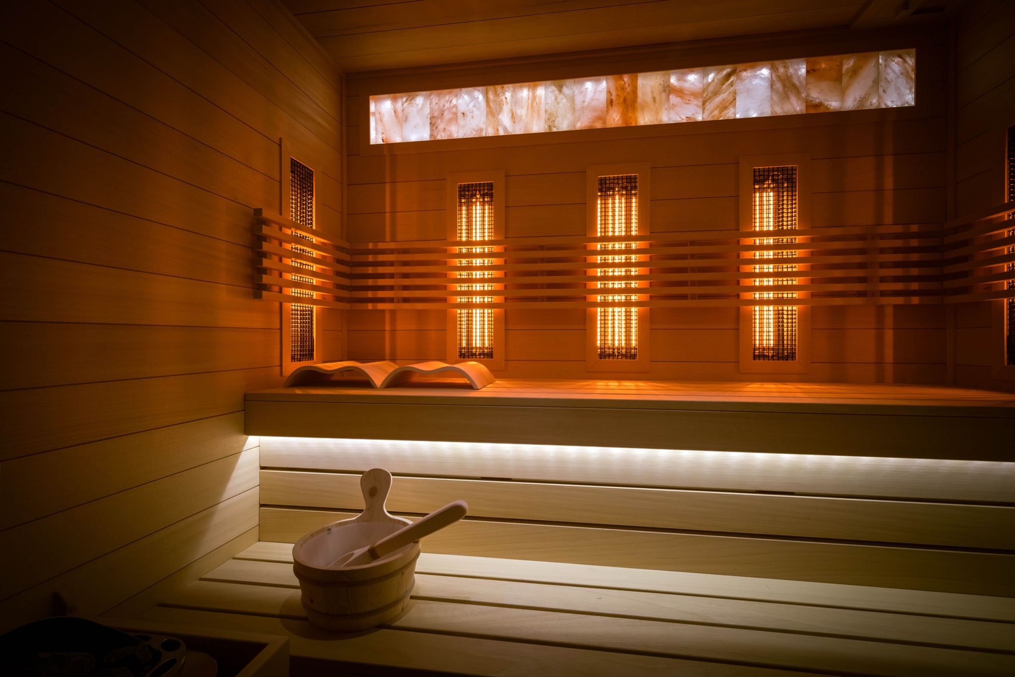 Wnętrze drewnianej kabiny z trzema grzejnikami w obudowach, oświetlonym panelem solnym i drewnianym wiadrem z chochlą na ławce, obok leży otwarta książka.