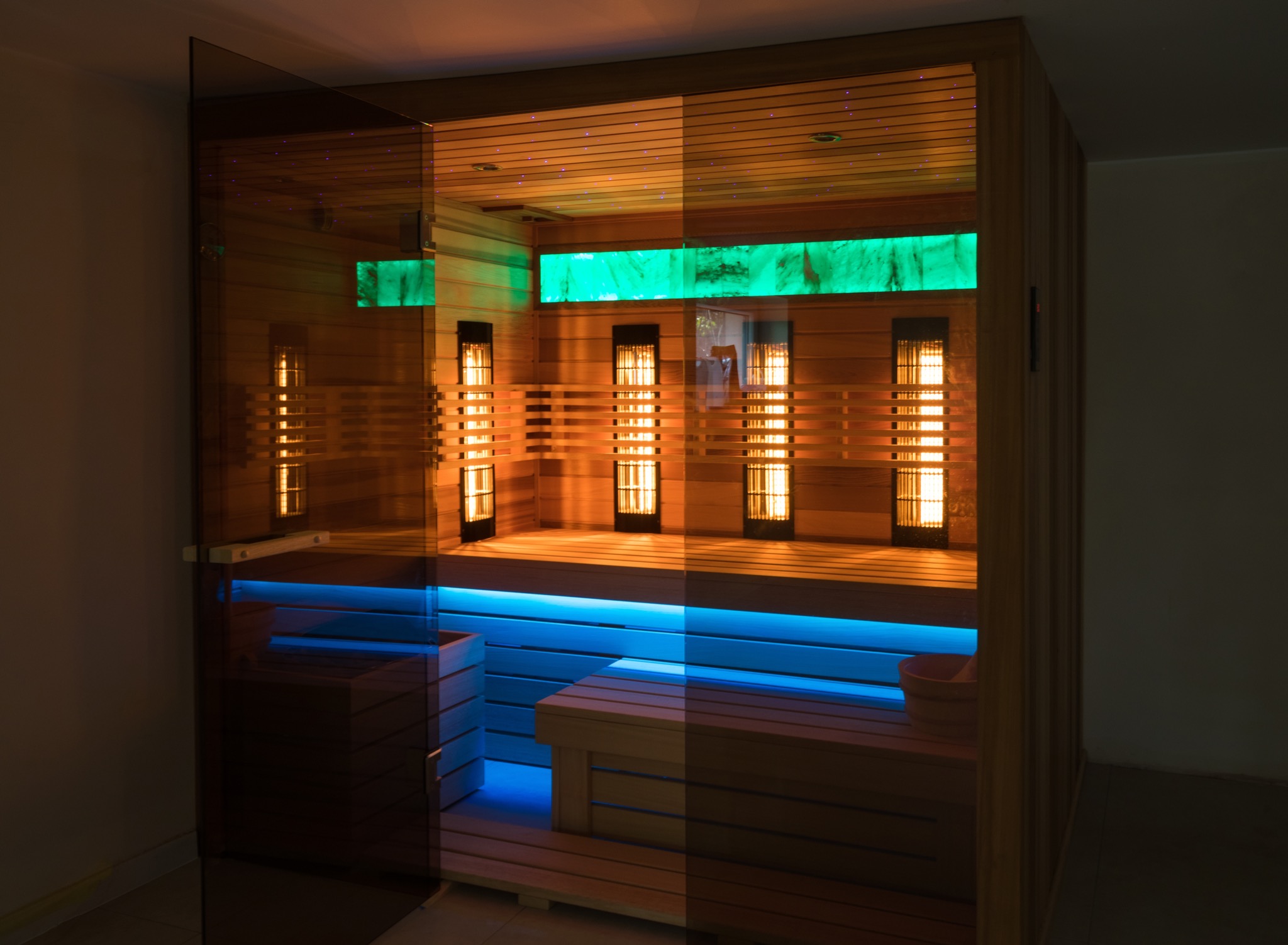 Nowoczesna kabina z drewna z podświetleniem LED w kolorach niebieskim i zielonym, oraz panelami grzewczymi. Szklane drzwi częściowo odbijają wnętrze.
