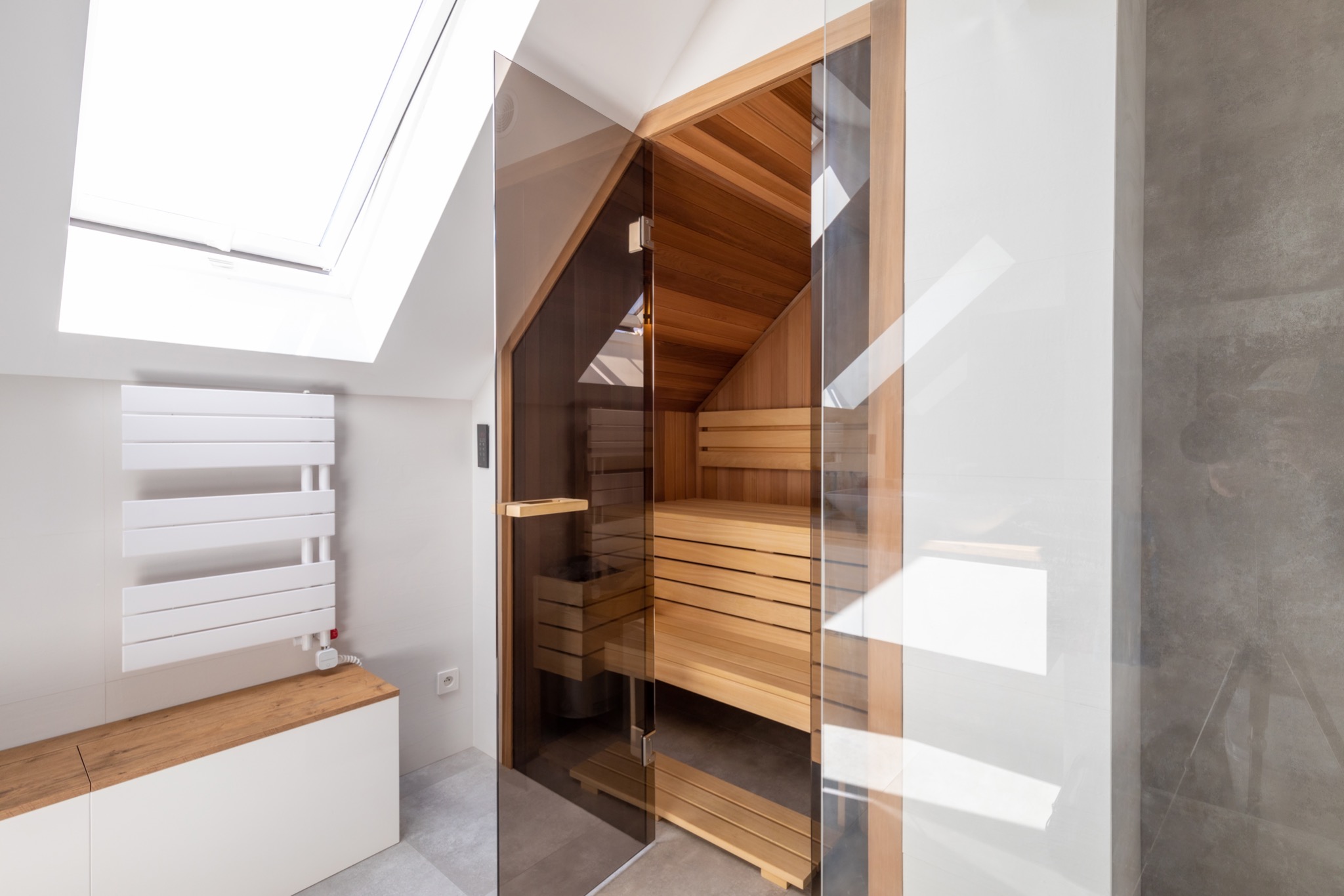 Nowoczesna sauna z przeszklonymi drzwiami, drewnianym wnętrzem i grzejnikiem drabinkowym w jasnej łazience z oknem dachowym.