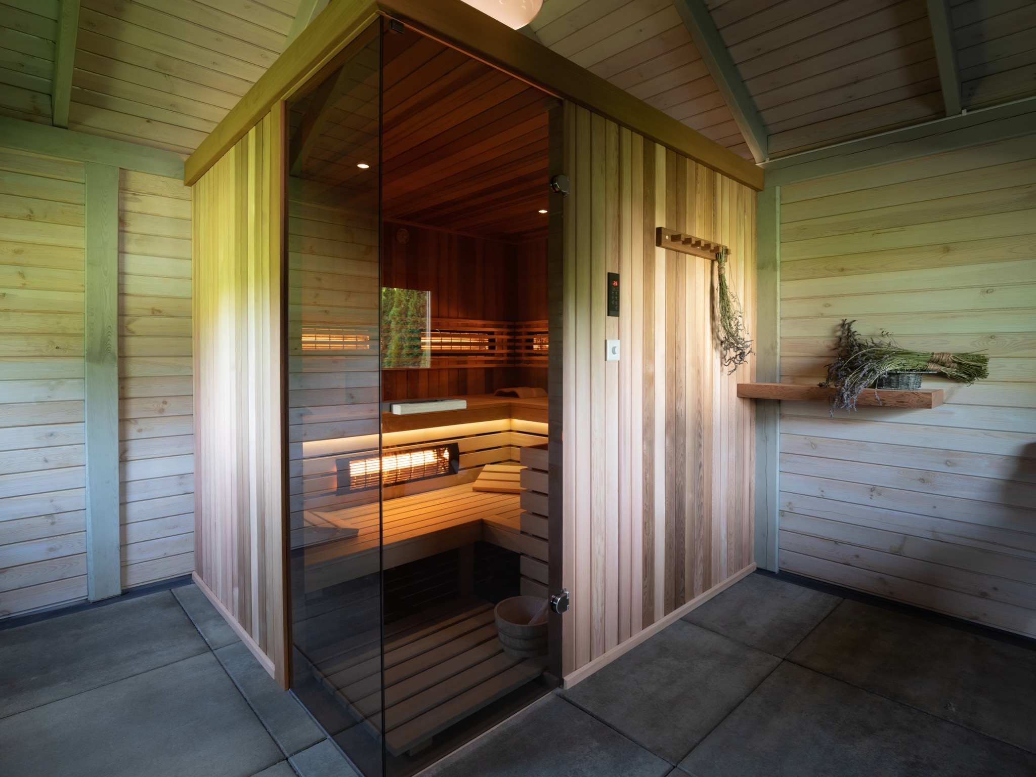 Drewniana kabina z przeszklonymi drzwiami, widoczne wnętrze z ławkami, piecem i drewnianym wiadrem, na ścianie półka z suszonymi ziołami.