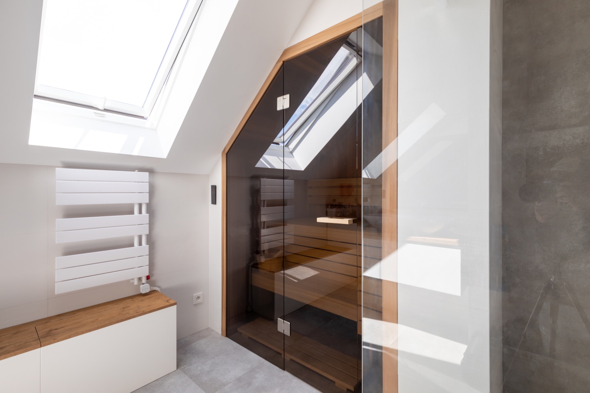 Nowoczesna kabina z drewna z przeszklonymi drzwiami, widok na wnętrze z drewnianymi ławkami, oświetlona światłem wpadającym przez okno dachowe, obok biały grzejnik drabinkowy i szara ściana.