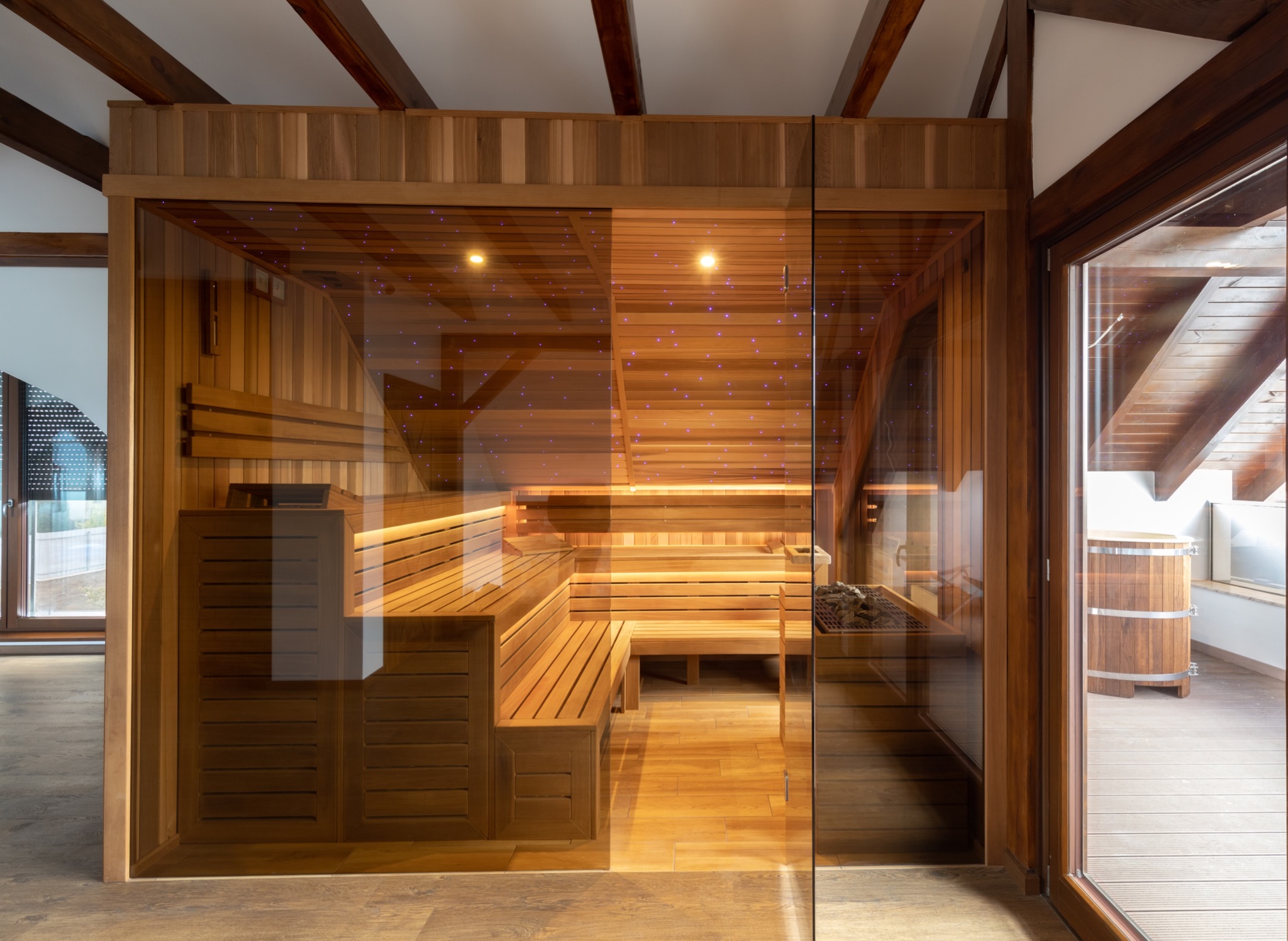 Nowoczesna, drewniana kabina z podświetleniem LED na suficie, widoczna przez szklaną ścianę. Obok kabiny stoi drewniana beczka z metalowymi obręczami.