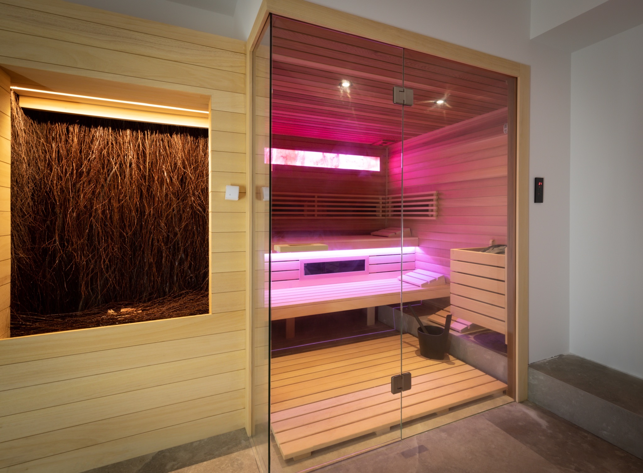 Nowoczesna kabina z drewna z przeszklonymi drzwiami, oświetlona różowym światłem, obok dekoracyjny panel z gałęzi.