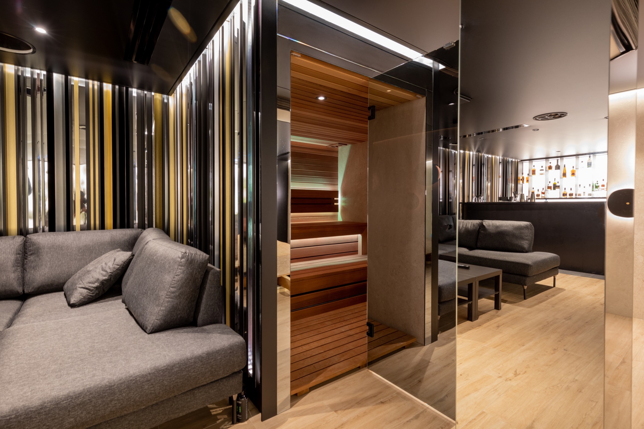 Luksusowe wnętrze z drewnianą kabiną, szarą sofą i barem w tle, całość odbija się w lustrzanych ścianach.