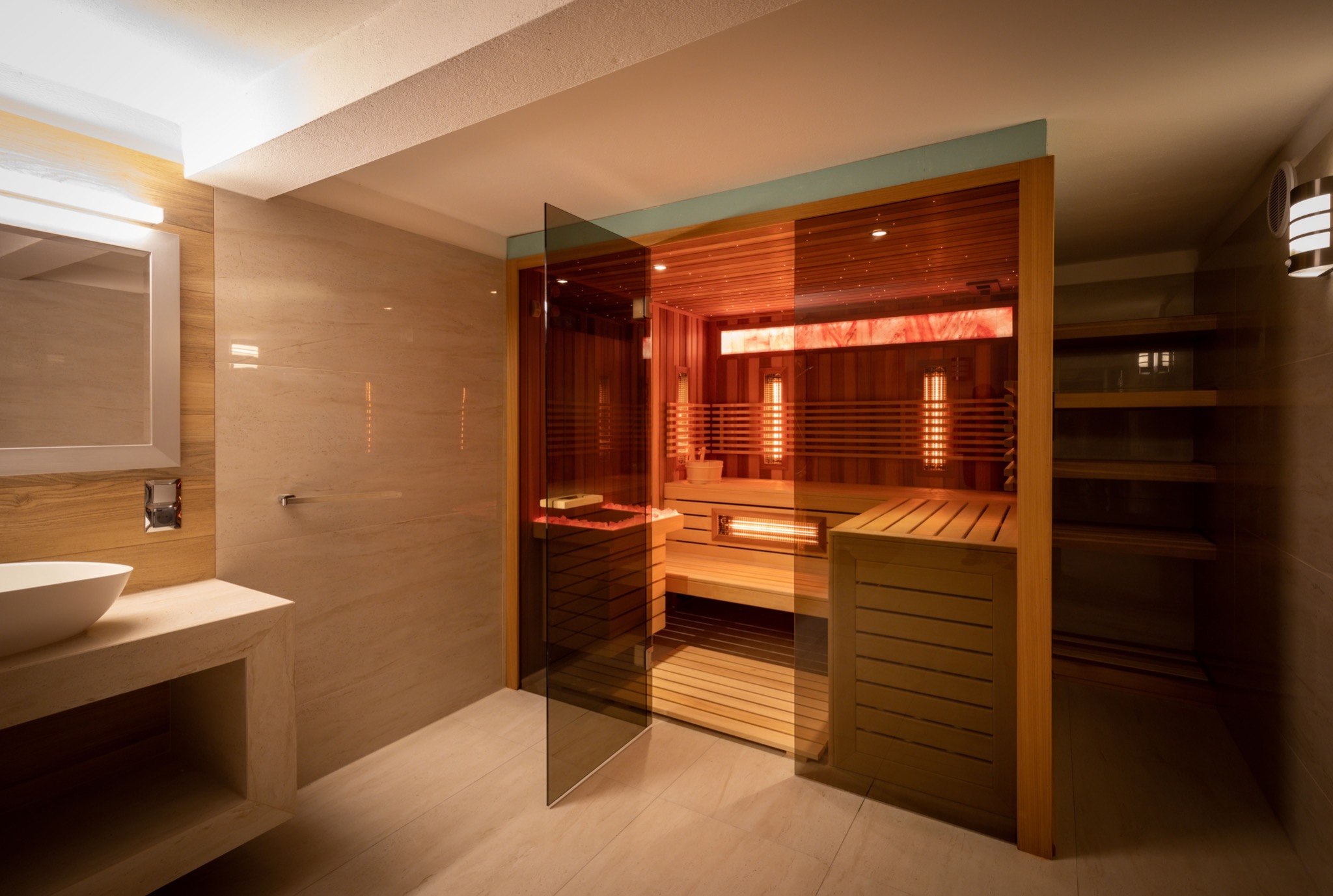 Nowoczesna sauna w łazience wykończonej w jasnych beżach, z przeszklonymi drzwiami, drewnianymi ławkami i panelami grzewczymi, oświetlona ciepłym światłem.