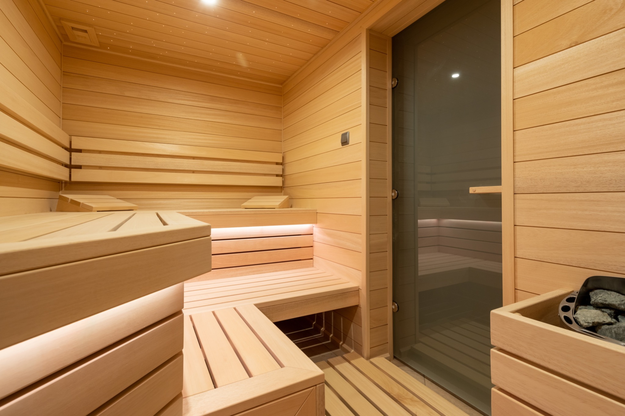 Jasne, drewniane wnętrze sauny z podświetlanymi ławkami, szklanymi drzwiami i kamieniami w pojemniku.