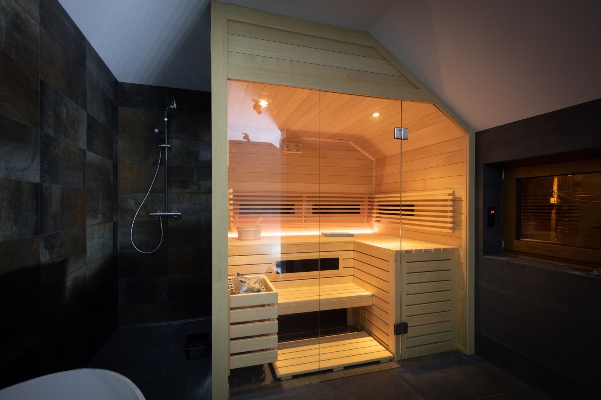 Nowoczesna drewniana kabina z przeszklonymi drzwiami, zlokalizowana w łazience z ciemnymi płytkami i prysznicem, wnętrze oświetlone ciepłym światłem.