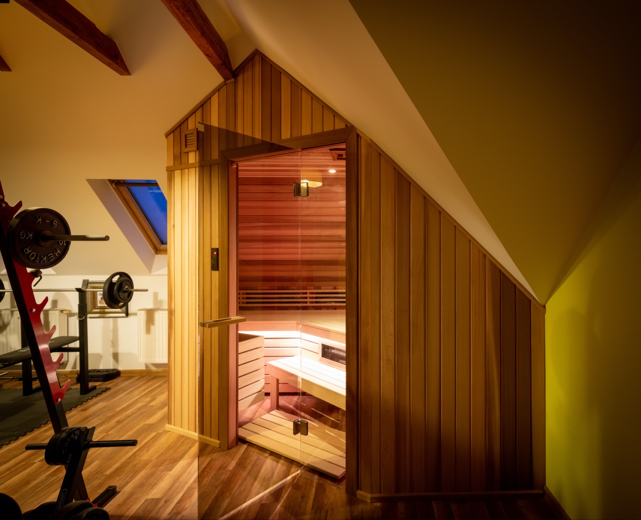 Drewniana kabina z przeszklonymi drzwiami w pomieszczeniu na poddaszu z oknem dachowym i sprzętem do ćwiczeń.