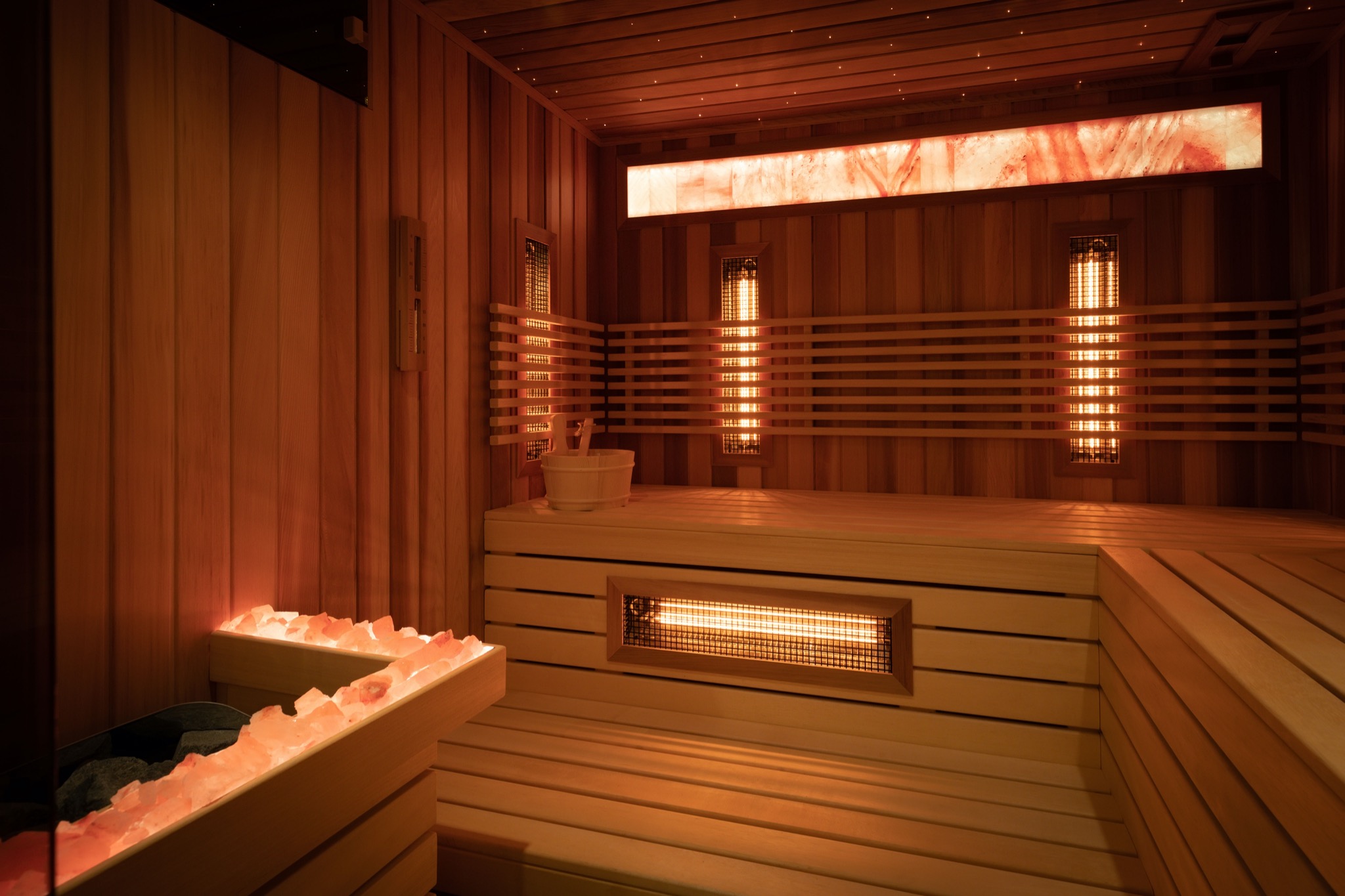 Wnętrze drewnianej kabiny z podświetlanymi panelami solnymi, piecem i akcesoriami do aromaterapii, oświetlone ciepłym światłem.