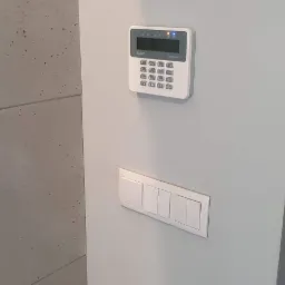 Panel sterowania alarmem domowym marki Satel Perfecto, zamontowany na szarej ścianie obok włączników światła w Zabrzu.