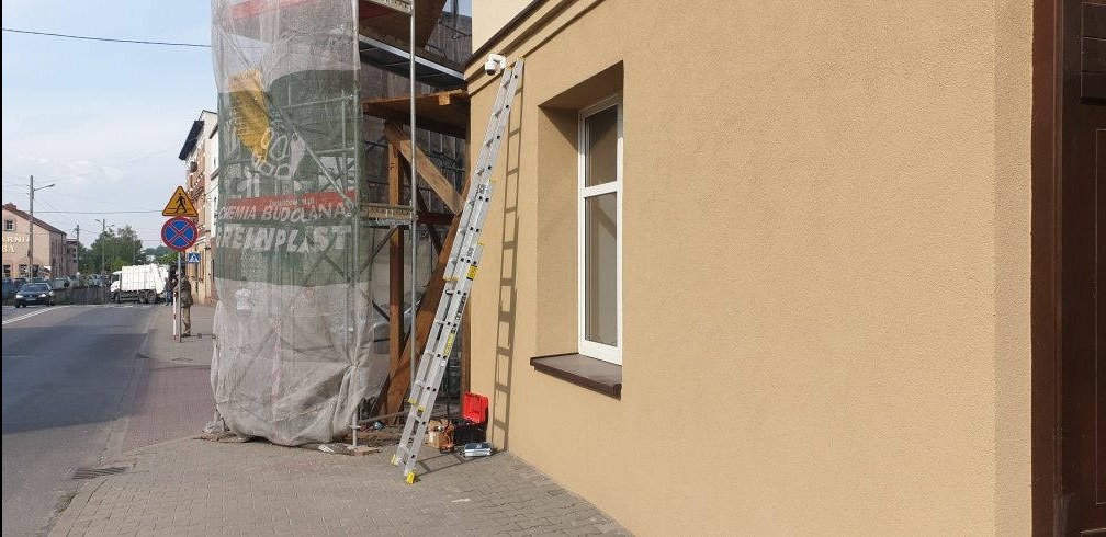 Kamera monitoringu zamontowana na elewacji budynku obok okna, obok drabina oparta o ścianę, w tle rusztowanie pod siatką ochronną.