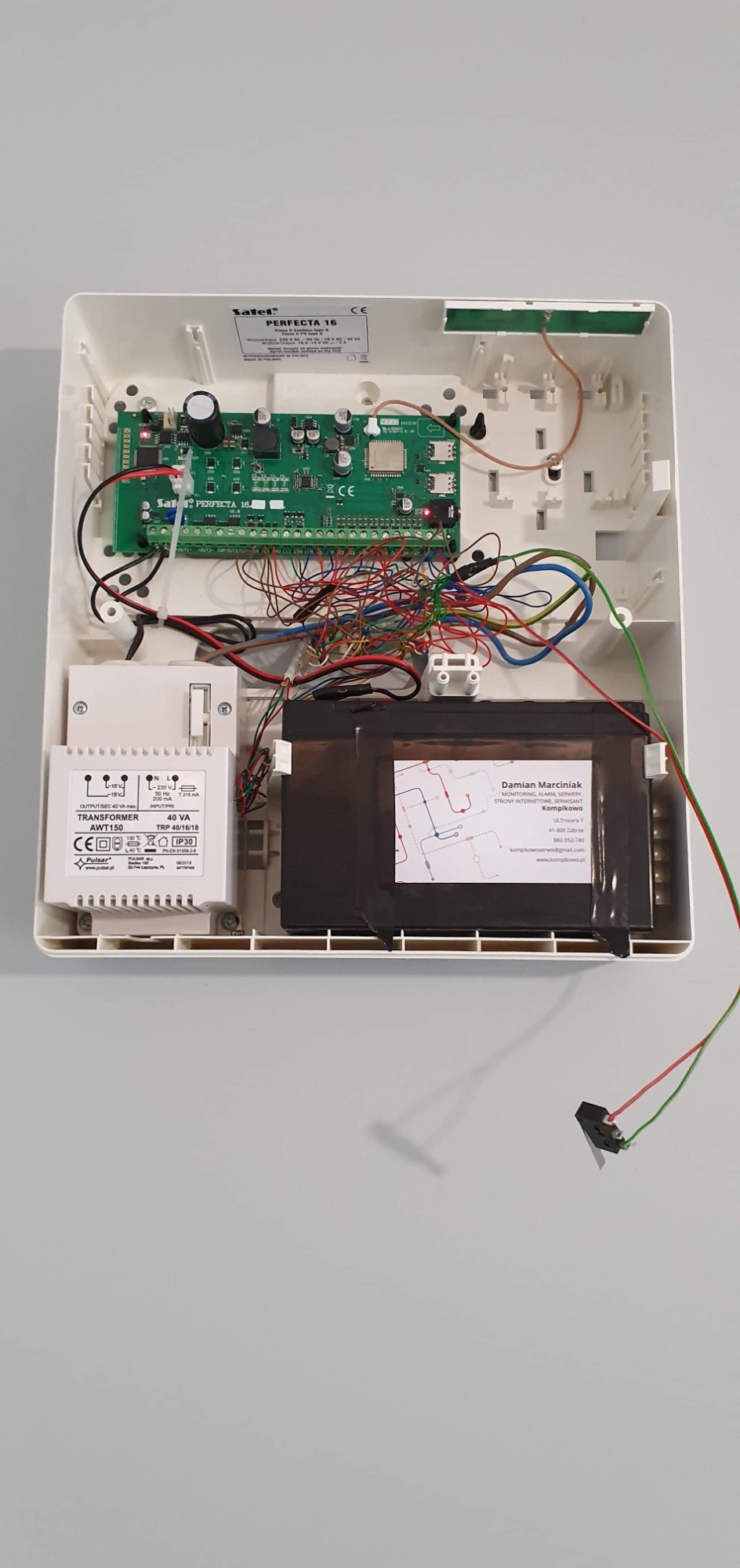 Otwarta obudowa centrali alarmowej Satel Perfecta 16 z widoczną płytą główną, transformatorem AWT150, akumulatorem podtrzymującym i plątaniną przewodów. Na akumulatorze naklejona wizytówka firmy...