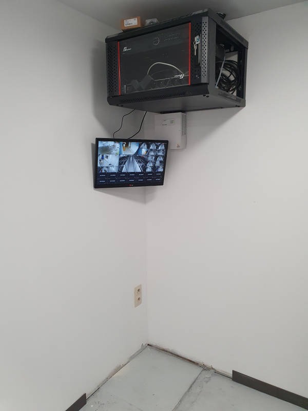 Zamontowany system monitoringu wizyjnego z ekranem pokazującym podgląd z wielu kamer, umieszczony w rogu pomieszczenia z widoczną instalacją elektryczną i szafką na sprzęt.