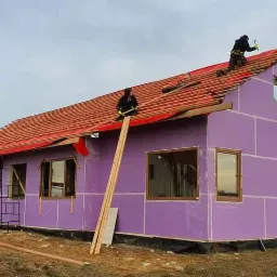 Budowa domu jednorodzinnego: montaż dachówki na czerwonej membranie, ściany zewnętrzne pokryte fioletowymi płytami izolacyjnymi, widoczni dwaj pracownicy na dachu, jeden z młotkiem, w tle inne...