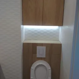 Nowoczesna toaleta podwieszana z zabudową w odcieniach drewna i oświetleniem LED, obok ściana wyłożona białymi płytkami z delikatnym wzorem.